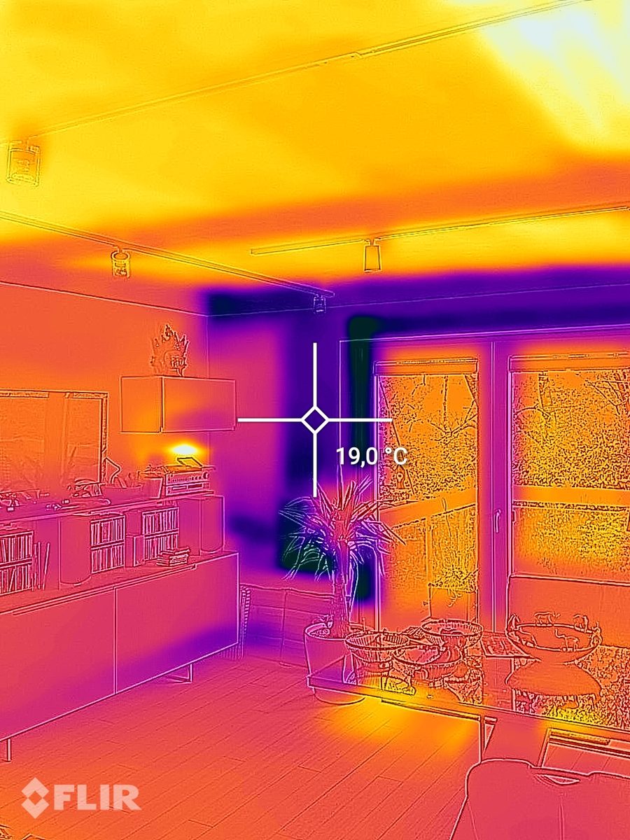 Imagem termográfica de uma sala de estar com teto quente devido ao aquecimento de teto e um ponto de medição de 19,0 graus na parede exterior junto à porta do terraço.